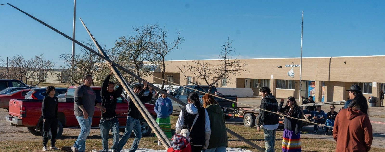 Several people from the school community stand around a tall pole, helping to construct a tipi in the schoolyard.