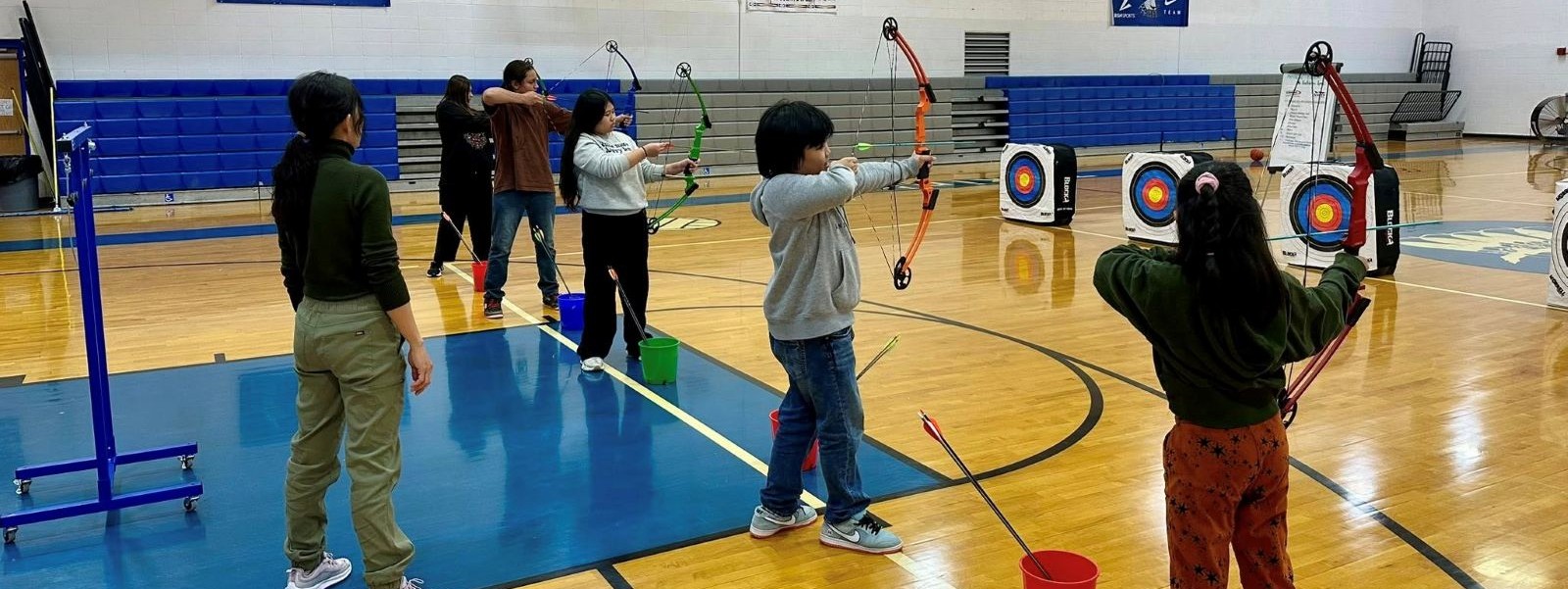 Students practicing archery indoors in the gym, focusing on their aim and technique.