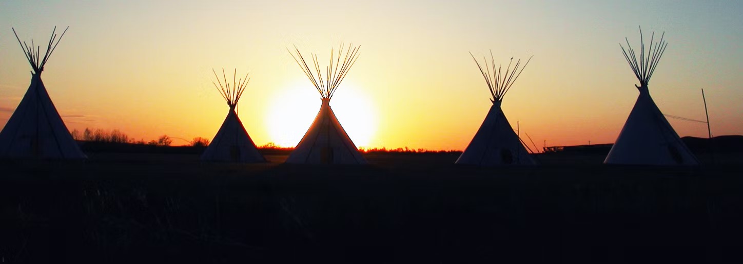 Several teepees stand together as the sun sets, casting a warm glow around them.