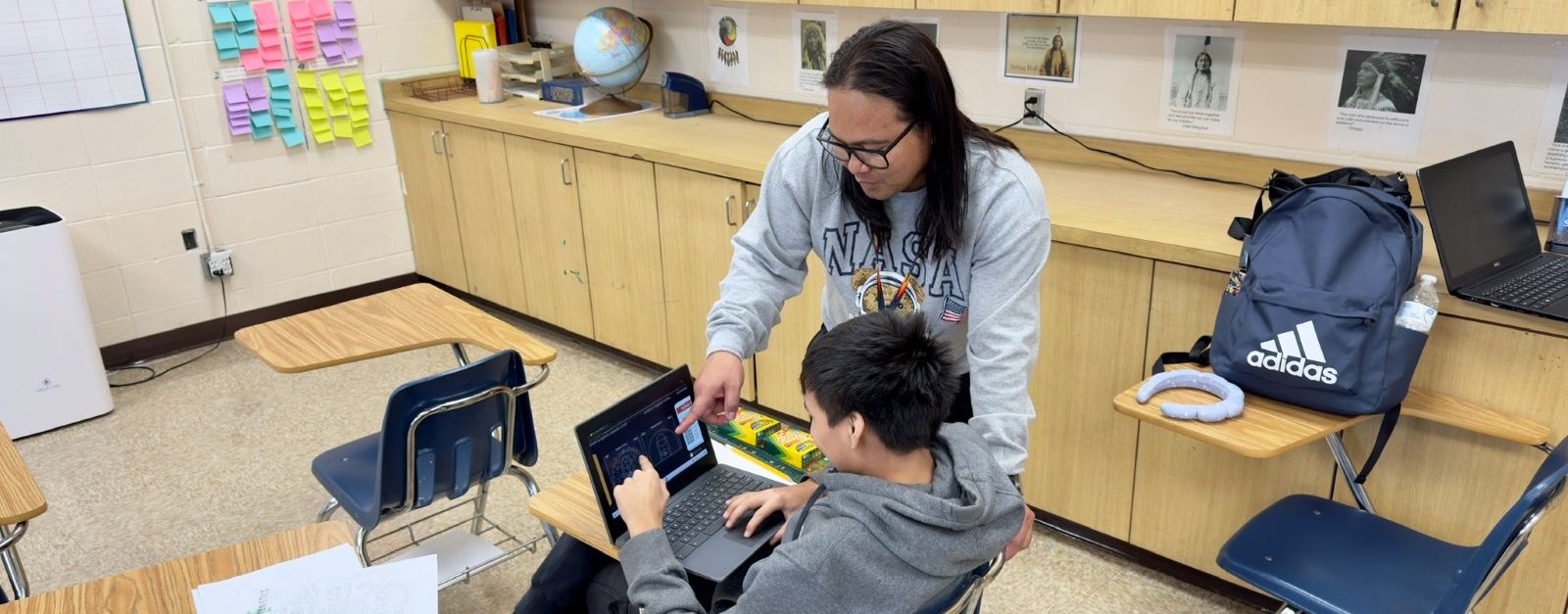 In a classroom, a teacher helps a student as they work together on a laptop, surrounded by learning materials.