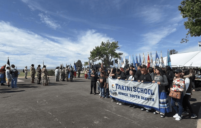 A group of people stands outdoors holding a banner for Takini School in Howes, South Dakota, with rows of flags and uniformed participants in the background.
