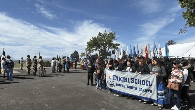 A group of people stands outdoors holding a banner for Takini School in Howes, South Dakota, with rows of flags and uniformed participants in the background.